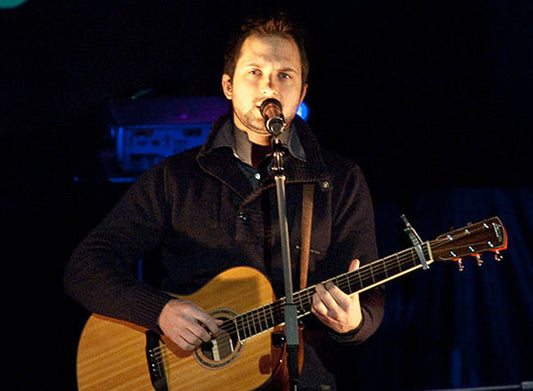 Male musician performing on stage with an acoustic guitar and microphone, captured during a live music performance with stage lighting in the background.