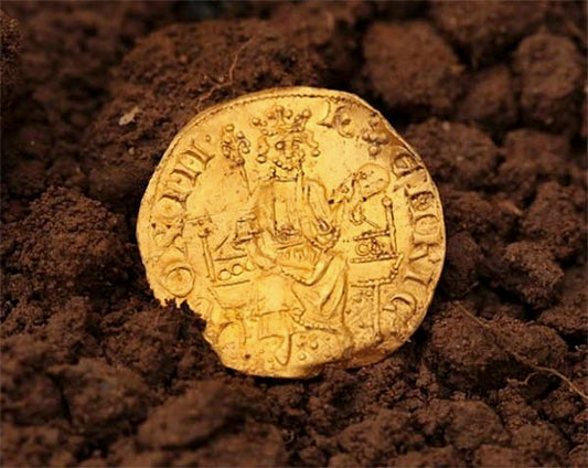 Close-up of a medieval gold coin buried in dirt, featuring an image of a crowned king seated on a throne. The coin is partially visible, showcasing its detailed engraving and historical significance.