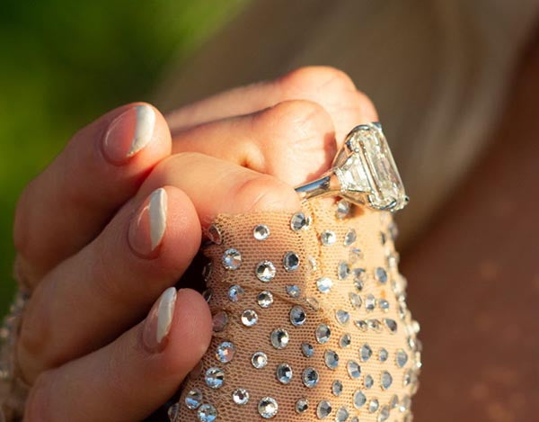 Close-up of a woman's hand with a large emerald-cut diamond engagement ring, adorned with sparkling rhinestones on a beige fabric background, highlighting the diamond's clarity and elegance.