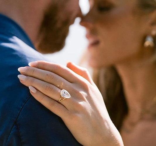 Close-up of a woman wearing a stunning pear-shaped diamond engagement ring, with her hand resting on a man’s shoulder as they share a romantic moment.