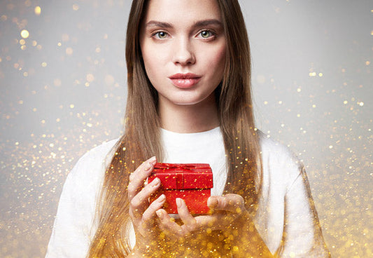 Young woman holding a red gift box with sparkling golden lights in the background.