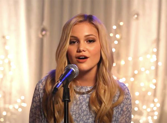 A young woman with long blonde hair singing into a microphone with twinkling lights in the background.