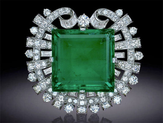 Elegant emerald brooch surrounded by sparkling diamonds, featuring a large square green emerald at the center with intricate diamond accents and swirling diamond ribbons.