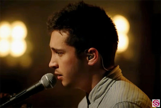 Profile shot of a male singer wearing an ear monitor and singing into a microphone with bright stage lights in the background.