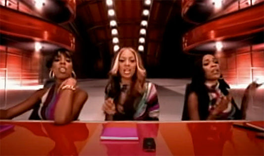 Three women performing together at a table in a studio, dressed in colorful outfits with bright lighting in the background.