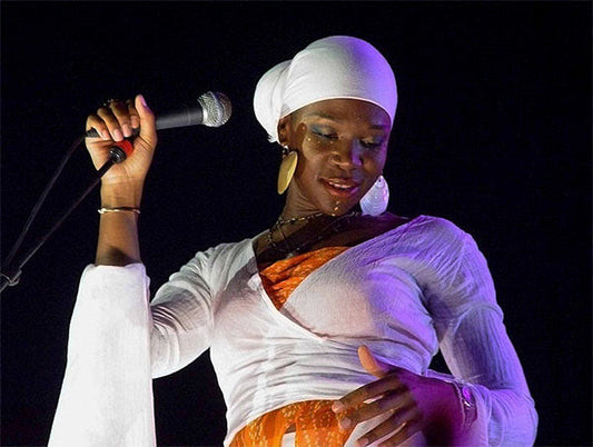Female singer performing on stage with a microphone, wearing a white headscarf, earrings, and a vibrant orange and white outfit.