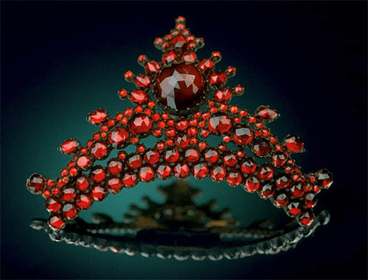 Close-up of a luxurious red gemstone tiara featuring a central large stone and smaller red gems arranged in an intricate pattern, set against a dark background.