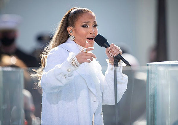 Jennifer Lopez performing at a public event, wearing a white coat and large earrings while singing into a microphone, with a confident expression.
