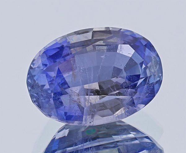Close-up of a large, oval-cut sapphire gemstone with a stunning violet-blue color. The gemstone's facets are clearly visible, reflecting light and showing its brilliance on a smooth reflective surface.