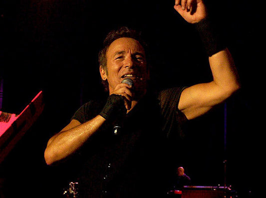 Bruce Springsteen performing live on stage, singing into a microphone with his arm raised during a concert.