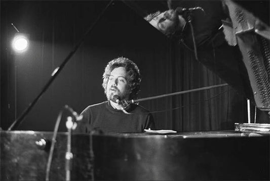 Black and white photo of a man performing at a piano on stage, with a microphone in front and his reflection visible in the piano's lid.