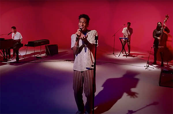 Jon Batiste performing live on stage with his band, surrounded by instruments like a piano, bass, and keyboard, against a vibrant red background.