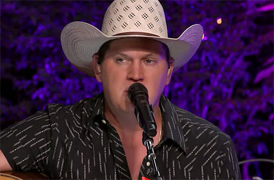 A man wearing a cowboy hat, singing into a microphone with a guitar in hand, performing on stage with purple lighting in the background.