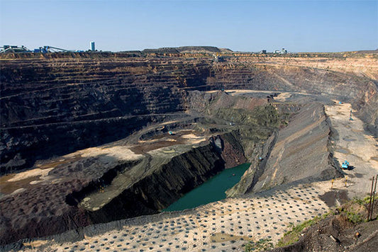Wide-angle view of an open-pit mining site with visible excavation, layers of earth, and a small body of water at the bottom of the pit.
