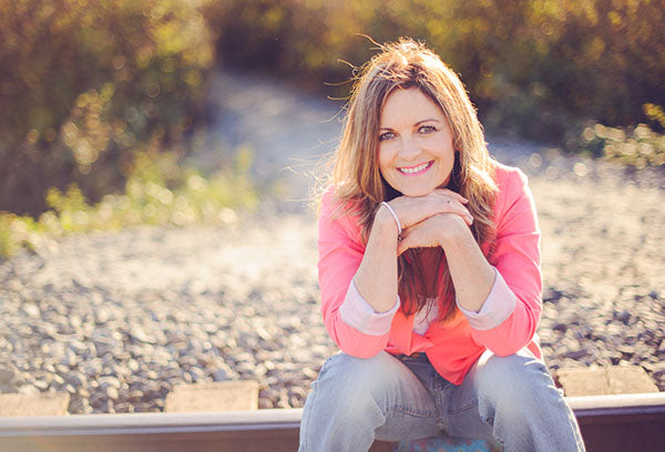 Smiling woman in a pink jacket sitting on railroad tracks, resting her chin on her hands with a sunny outdoor background.