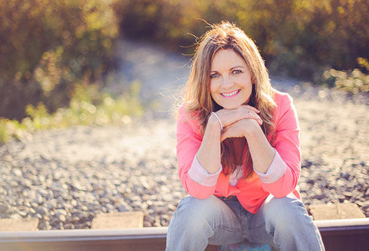 Smiling woman in a pink jacket sitting on railroad tracks, resting her chin on her hands with a sunny outdoor background.