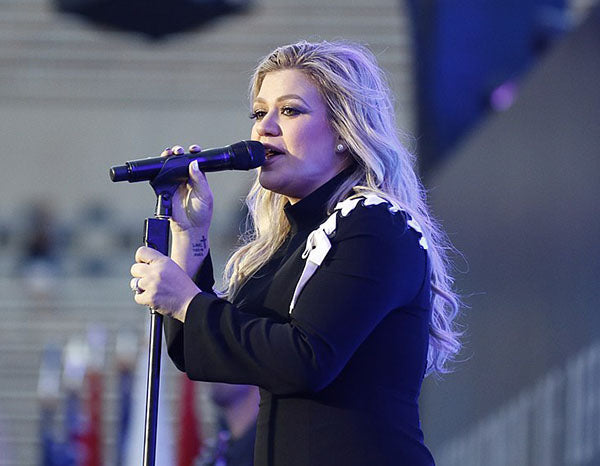 Kelly Clarkson performing live on stage, singing into a microphone with blonde hair styled in waves, wearing a black dress with white accents.