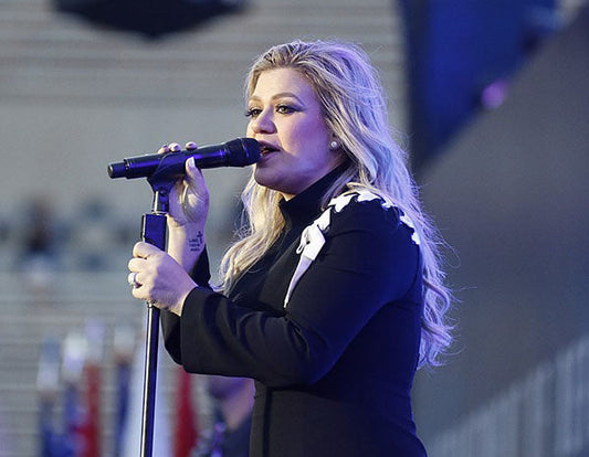 Kelly Clarkson performing live on stage, singing into a microphone with blonde hair styled in waves, wearing a black dress with white accents.