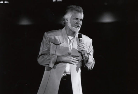 Kenny Rogers performing on stage with a microphone, wearing a light-colored jacket and a white shirt, black and white photo.