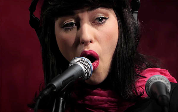 Close-up of a female singer performing into a microphone, wearing headphones. She has bold red lipstick, winged eyeliner, and a pink scarf around her neck, with a focused expression as she sings in front of a red background.