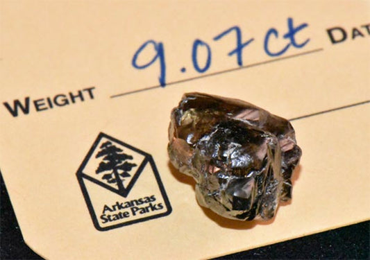 Close-up of a rough diamond with a weight of 9.07 carats, placed on an Arkansas State Parks label. The diamond's natural form is visible, with a yellow background highlighting its details.