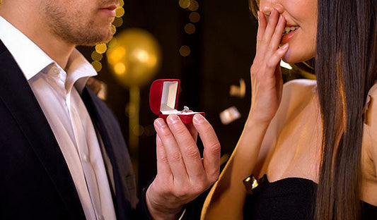 Man proposing with an engagement ring inside a red velvet box, while woman reacts with excitement and covers her mouth in surprise at the proposal.