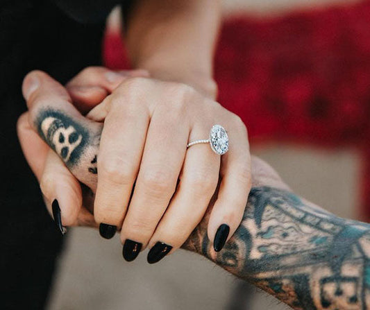 Close-up image of an engagement ring on a hand with black nails, showcasing a large oval diamond ring, paired with a tattooed hand. The couple's hands are intertwined, symbolizing love and commitment.