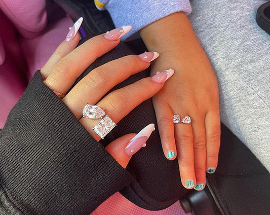 Close-up of two hands with manicured nails, featuring elegant rings on each hand, one with a large diamond and the other with smaller stones, set against a soft pink background.