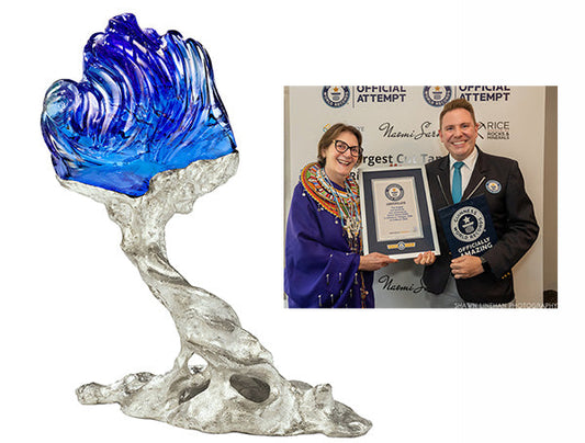 Sculpture of a wave in blue and clear glass, symbolizing the largest cut crystal wave, displayed alongside a photo of a man and a woman holding a Guinness World Records certificate for the largest crystal wave.