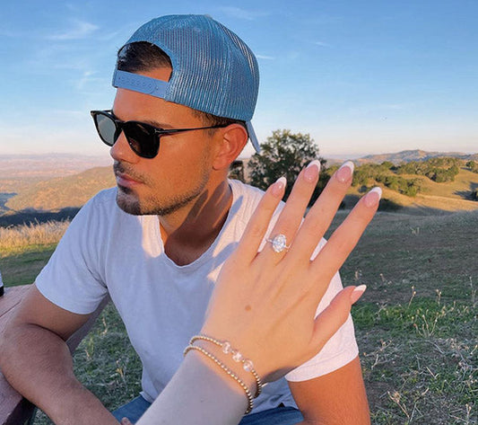 Engagement photo of a woman showing off her sparkling diamond ring while seated next to a man wearing sunglasses and a blue cap, with a scenic outdoor backdrop.