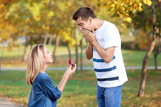 Woman proposing with an engagement ring to a surprised man in a park during the fall, surrounded by colorful autumn leaves.