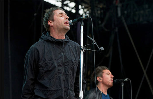 Liam Gallagher performing on stage with his brother Noel Gallagher in the background, during an Oasis concert.