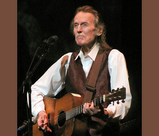 Legendary musician Gordon Lightfoot performing live on stage with his guitar, wearing a vest and shirt, singing passionately into the microphone.