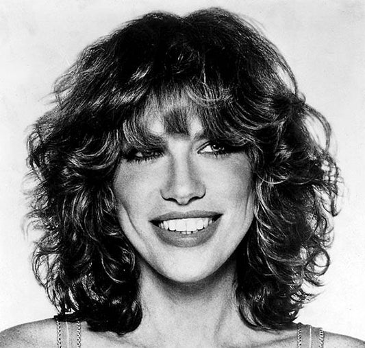 Black and white portrait of a smiling woman with voluminous curly hair and bangs. The image captures her bright expression and natural beauty.