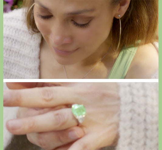 Close-up image of a woman with hoop earrings, showing an emotional expression while admiring a green gemstone ring with diamond accents.
