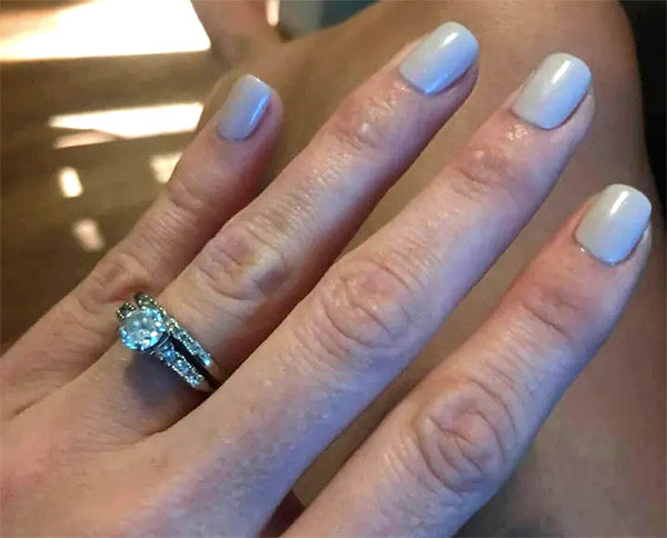 Close-up of a hand showing an engagement ring and wedding band with a diamond center stone, with light beige nail polish on fingers.