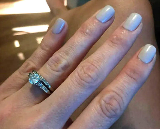 Close-up of a hand showing an engagement ring and wedding band with a diamond center stone, with light beige nail polish on fingers.