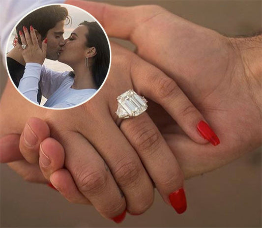 Close-up of an engagement ring on a couple's hands, featuring a large emerald-cut diamond. The woman’s red nails highlight the ring, and the background shows a romantic kiss with the couple in the inset.