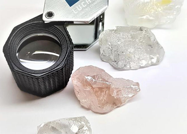 A jeweler's loupe and several rough diamond stones, including clear and pink diamonds, placed on a white surface.