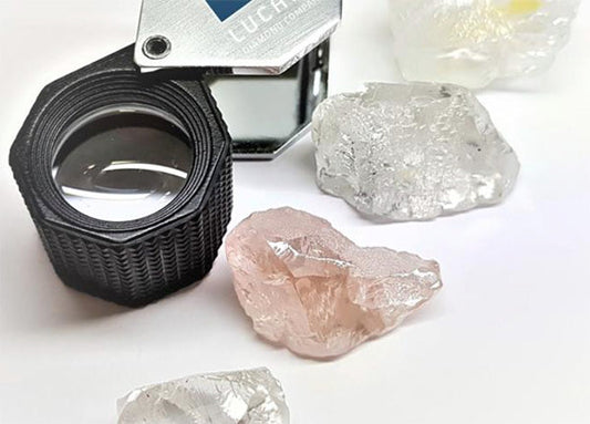 A jeweler's loupe and several rough diamond stones, including clear and pink diamonds, placed on a white surface.