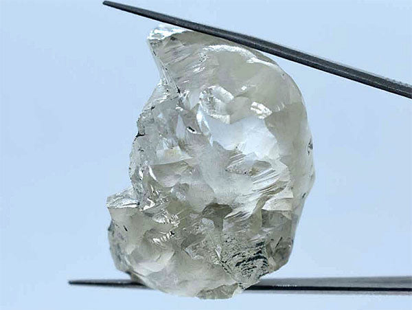 A close-up image of a rough diamond held with tweezers, showcasing its raw, uncut texture and clarity.