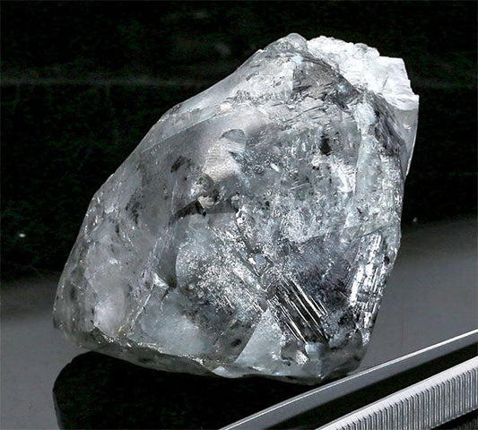 Close-up image of a rough diamond crystal with visible natural inclusions and texture, resting on a reflective surface with a tweezer beside it.