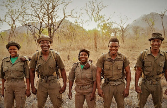 A group of five park rangers standing together in the African wilderness, smiling and dressed in khaki uniforms with backpacks.