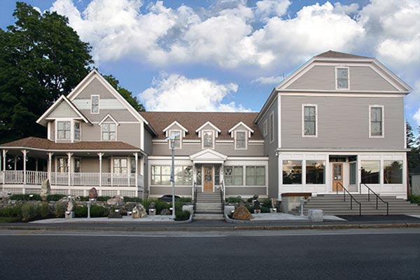 Exterior view of a large, two-story gray house with a front porch, modern entrance, and landscaped garden, under a partly cloudy sky.