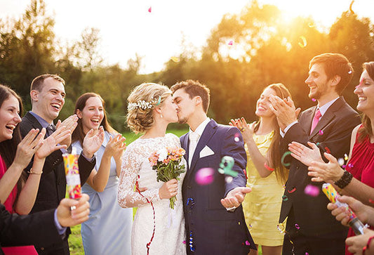 A newlywed couple sharing a kiss surrounded by friends and family at an outdoor wedding, with confetti flying in the air and everyone smiling and celebrating under the warm glow of the sunset.