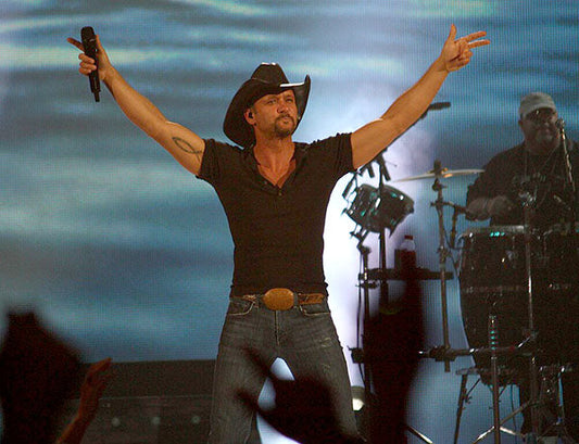 Tim McGraw performing on stage with his arms raised, wearing a cowboy hat and black shirt during a live concert.