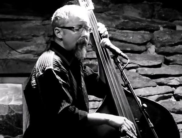 Black and white photo of a male jazz musician playing the double bass on stage. He is wearing glasses and a black shirt, with a microphone in front of him and a stone wall backdrop.