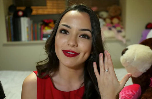 Woman showing off her engagement ring and smiling, wearing red lipstick, with a cozy background featuring stuffed animals and books.