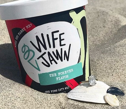 Ice cream tub labeled 'Wife Jawn The Forever Flavor' with a diamond ring and seashells on the sandy beach, symbolizing a proposal.