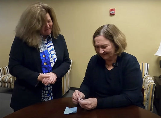 Two women in an office setting, one smiling while holding a ring and the other watching closely, likely during a meaningful moment of discovery or gift exchange.